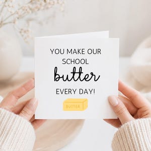 May include: A white greeting card with the text "YOU MAKE OUR SCHOOL butter EVERY DAY!" in black script. A yellow butter stick illustration is at the bottom. The card is held by two hands.