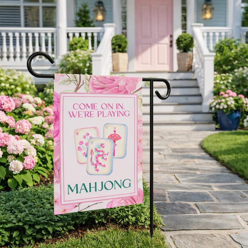 May include: A pink and white garden flag with the text "COME ON IN, WE'RE PLAYING MAHONG". The flag features a floral design and illustrations of Mahjong tiles. The flag is displayed on a black metal stand in a garden setting.