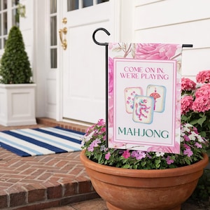 May include: A garden flag with a floral design and the words "Come on in, We're Playing Mahjong." The flag features three Mahjong tiles with colorful illustrations. The flag is displayed in a flower pot filled with pink flowers.