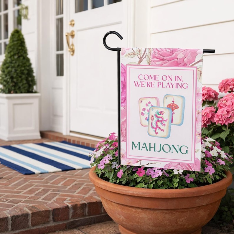 May include: A garden flag with a floral design and the words "Come on in, We're Playing Mahjong." The flag features three Mahjong tiles with colorful illustrations. The flag is displayed in a flower pot filled with pink flowers.