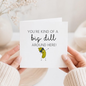 May include: A white greeting card with the text "YOU'RE KIND OF A big dill AROUND HERE!" and a cartoon pickle wearing sunglasses. The card is held by two hands, with a neutral background.