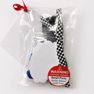Puede incluir: Una bolsa de pl&aacute;stico transparente atada con una cinta roja, que contiene varios materiales de manualidades. Incluye una forma blanca con bordes festoneados, una cinta a cuadros en blanco y negro y una etiqueta de advertencia: "Peligro de asfixia, piezas peque&ntilde;as, no apto para ni&ntilde;os menores de 3 a&ntilde;os."
