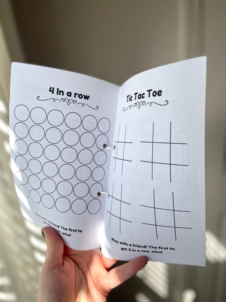 K&ouml;nnte beinhalten: Ein schwarz-wei&szlig;es druckbares Spielheft mit zwei Spielen: 4 in einer Reihe und Tic Tac Toe. Das Heft ist auf der Tic Tac Toe-Seite ge&ouml;ffnet, die ein Gitter mit Linien f&uuml;r die Spieler zum Markieren ihrer Felder hat.