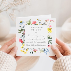 May include: A white card featuring the words "THINKING OF YOU" and a cross, framed by a border of vibrant wildflowers. The card also has a message. The card is held by a person.