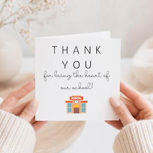 Pode incluir: Um cartão branco com as palavras "THANK YOU" em preto, seguido da frase manuscrita "for being the heart of our school!". Uma ilustração colorida de uma escola está na parte inferior.