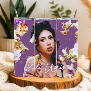 May include: A set of three stainless steel tumblers featuring a portrait of Kali Uchis against a purple background. The tumblers are adorned with floral accents and the artist's name in a cursive font.