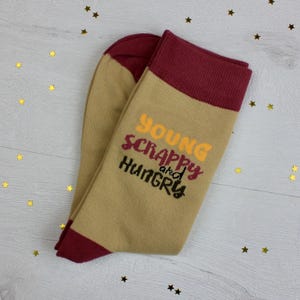 May include: A pair of tan socks with claret red accents on the toe and cuff. The socks feature the text "Young, Scrappy, and Hungry" in yellow, claret red, and black lettering. The socks are on a white wooden surface with gold star-shaped confetti.