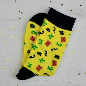 May include: A pair of yellow socks with black heels and cuffs. The socks feature a pattern of green, black, and red icons, including question marks, briefcases, and envelopes. The socks are on a white wooden surface.