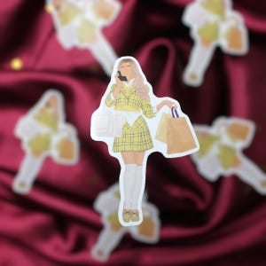 May include: A sticker featuring a cartoon illustration of a woman in a yellow plaid outfit, holding shopping bags and a phone to her ear. The sticker has a white border and is set against a burgundy background.