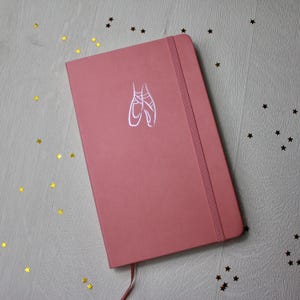 May include: A dusky pink notebook featuring a ballet slipper design. The notebook has an elastic band closure and a ribbon bookmark. Gold star-shaped confetti is scattered around the notebook.