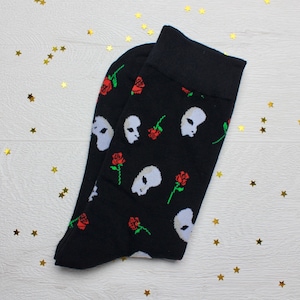 May include: Black socks featuring a repeating pattern of white alien faces and red roses. The socks have a black cuff. The socks are displayed on a white surface with gold star-shaped confetti.
