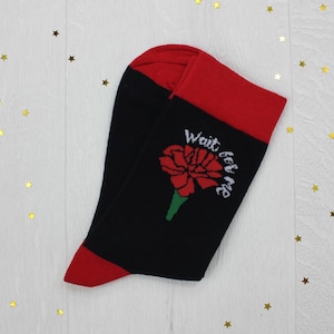 May include: A pair of black crew socks featuring a red carnation flower graphic with a green stem and the white text "Wait for me" printed above the flower. The socks have a solid red cuff at the top and a solid red toe section at the bottom.