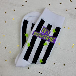 May include: White socks with black vertical stripes and the text "IT'S SHOWTIME" in green and purple. The socks also feature green splatters. The socks are on a light wood surface with gold star decorations.