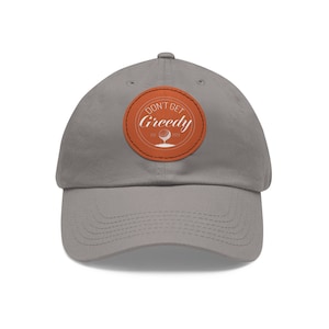 Könnte beinhalten: Eine graue Baseballkappe mit einem braunen Lederpatch, auf dem "Don't Get Greedy" mit einer Golfballgrafik steht.