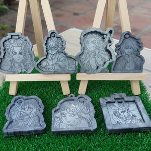 Könnte beinhalten: Sechs graue Schlüsselanhänger aus Kunstharz mit detaillierten Anime-Charakterporträts. Jeder Schlüsselanhänger hat ein einzigartiges Design mit sichtbaren Gesichtszügen und Kleidungsdetails. Präsentiert auf einer grünen Oberfläche mit kleinen Holzstaffeleien.