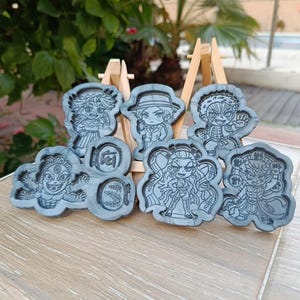 Könnte beinhalten: Eine Sammlung grauer, harzgeformter Charaktermolden, jede mit detaillierten eingravierten Designs. Die Formen werden auf einer hölzernen Staffelei präsentiert und zeigen verschiedene Figuren im Anime-Stil. Der Hintergrund zeigt Grün und eine Holzoberfläche.