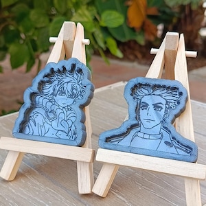 Könnte beinhalten: Zwei graue Kunstwerke aus Kunstharz mit Anime-Charakteren, präsentiert auf kleinen Holzstaffeleien. Die Kunstwerke weisen detaillierte Strichzeichnungen auf und sind vor einem verschwommenen grünen Hintergrund platziert. Die Staffeleien sind aus hellem Holz gefertigt.