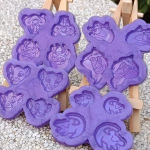 May include: Three purple silicone moulds featuring embossed character designs from The Lion King. Each mould contains multiple distinct impressions of various characters, including lions, a warthog, and a meerkat, with detailed outlines.