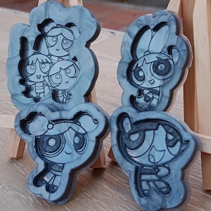 Könnte beinhalten: Vier graue Harzformen der Powerpuff Girls-Charaktere. Die Formen zeigen die Zeichentrickfiguren Blossom, Bubbles und Buttercup. Die Formen werden auf einer hölzernen Staffelei ausgestellt. Die Formen eignen sich zum Basteln.