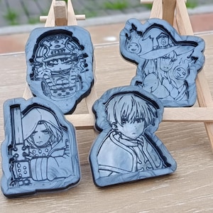 以下が含まれることがあります： 4つの灰色の樹脂製アニメキャラクターフィギュア。各フィギュアは異なるキャラクターで、詳細な線画が施されています。フィギュアは小さな木製のイーゼルに展示されています。フィギュアは様々な形をしています。