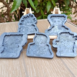 Könnte beinhalten: Fünf graue Resin-Formen mit Porträts von Anime-Charakteren. Jede Form zeigt einen anderen Charakter mit detaillierten Gesichtszügen und Frisuren. Die Formen werden auf kleinen Holzstaffeleien vor grünem Laubwerk präsentiert.