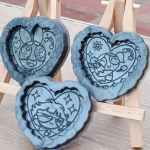 Könnte beinhalten: Drei herzförmige Formen mit Cartoon-Charakter-Designs. Die Formen sind marmoriert grau mit schwarzen Umrissen. Jedes Herz zeigt einen anderen Charakter mit floralen und Regenbogen-Akzenten. Die Formen werden auf kleinen Holzstaffeleien präsentiert.