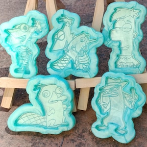 May include: Five teal-colored silicone molds featuring cartoon characters. The molds depict characters in various poses, with detailed outlines and a slightly translucent appearance. The molds are arranged on a small wooden easel.
