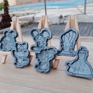 Könnte beinhalten: Eine Sammlung grauer Formen in Form von Cartoon-Charakteren, die auf kleinen Holzstaffeleien ausgestellt sind. Die Formen zeigen Charaktere wie Jerry, Courage, der feige Hund, und Samurai Jack. Der Text "MAMA ADKINS" ist auf einer Form zu sehen.