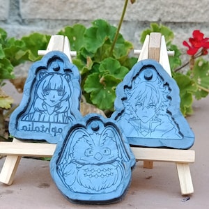 Könnte beinhalten: Drei blaue Resin-Anhänger mit Charakterdesigns, jeweils auf einem kleinen Holzstaffelei montiert. Die Anhänger zeigen detaillierte Illustrationen von Anime-Charakteren und einer Eule. Ein Anhänger hat arabischen Text.