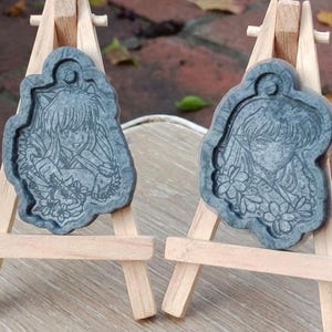 Könnte beinhalten: Zwei graue Resin-Anhänger mit detailliertem Anime-Charakter-Design, jeweils auf einer kleinen Holzstaffelei präsentiert. Die Anhänger weisen ein Blumenmotiv auf und sind vor einem verschwommenen Hintergrund im Freien platziert.