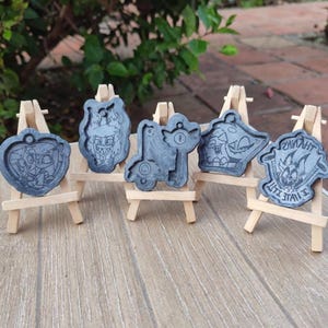 Könnte beinhalten: Sechs graue Resin-Anhänger mit Cartoon-Charakter-Designs, die auf kleinen Holzstaffeleien ausgestellt sind. Die Anhänger haben verschiedene Formen, darunter ein Herz, ein Schlüssel und ein Schild. Der Hintergrund ist verschwommen, mit grünem Laub sichtbar.