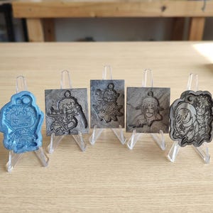 Puede incluir: Una colección de colgantes temáticos de personajes exhibidos en pequeños caballetes transparentes. Los colgantes presentan diseños detallados en varios colores, incluyendo azul y gris. Los colgantes son cuadrados y rectangulares, mostrando diferentes personajes.