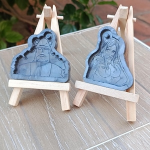 Puede incluir: Dos figuras grises de resina de personajes de anime, exhibidas en pequeños caballetes de madera. Las figuras están detalladas con líneas grabadas, mostrando los rasgos de los personajes. El fondo está borroso.