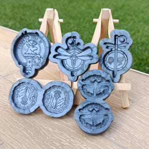 Könnte beinhalten: Eine Sammlung grauer Harzformen mit verschiedenen Designs, darunter ein Hirsch, eine Libelle und eine Distel. Die Formen werden auf einer kleinen Holzstaffelei vor dem Hintergrund von grünem Gras und einer Holzoberfläche präsentiert. Die Formen sind in verschiedenen Formen und Größen.