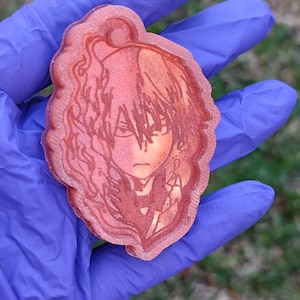 Könnte beinhalten: Ein roter Schlüsselanhänger aus Harz mit einer detaillierten Illustration eines Anime-Charakters mit schwarzen Haaren und einem ernsten Ausdruck. Der Charakter trägt ein dunkles Outfit mit einem weißen Kragen.