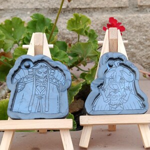 Könnte beinhalten: Zwei graue, wolkenförmige Anhänger mit detaillierten schwarzen Strichzeichnungen, die auf kleinen Holzstaffeleien ausgestellt sind. Einer zeigt eine skelettartige Figur, der andere eine Zeichentrickfigur. Der Hintergrund umfasst rote Blumen und grünes Laub.