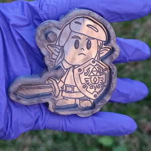 Könnte beinhalten: Ein Holzschlüsselanhänger mit einer Comicfigur, die ein Schwert und einen Schild hält. Die Figur trägt eine grüne Tunika und einen braunen Hut. Der Schild hat ein goldenes Design mit einem stilisierten Dreieck und einem Kreis.