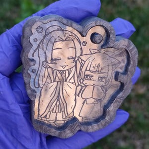 Könnte beinhalten: Ein Holzschlüsselanhänger mit einem lasergravierten Design von zwei Anime-Charakteren. Die Charaktere sind im Chibi-Stil gezeichnet und tragen traditionelle japanische Kleidung. Der Schlüsselanhänger ist braun mit einer dunklen Holzmaserung.