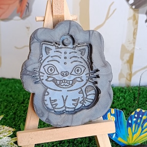 Könnte beinhalten: Grauer, katzenförmiger Anhänger mit detailliertem, cartoonartigem Design, ausgestellt auf einer kleinen Holzstaffelei. Die Katze hat große Augen und ein grinsendes Maul. Der Anhänger ist von einem dekorativen Rand umgeben.