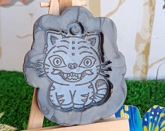 Könnte beinhalten: Grauer, katzenförmiger Anhänger mit detailliertem, cartoonartigem Design, ausgestellt auf einer kleinen Holzstaffelei. Die Katze hat große Augen und ein grinsendes Maul. Der Anhänger ist von einem dekorativen Rand umgeben.