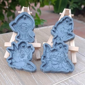 Könnte beinhalten: Vier graue Kunstharzformen von Zeichentrickfiguren werden auf kleinen Holzstaffeleien präsentiert. Die Formen weisen detaillierte Umrisse verschiedener Figuren auf, jede mit einzigartigen Designs. Der Hintergrund ist eine verschwommene Außenumgebung.