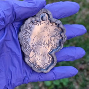 Könnte beinhalten: Ein Anhänger mit einem detaillierten Anime-Charakter-Design. Der Anhänger hat eine kupferfarbene Mitte mit einem detaillierten geätzten Bild eines Charakters, umgeben von einem grau-blauen Rand. Er hat oben eine kleine Schlaufe zur Befestigung.