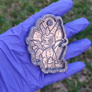 Könnte beinhalten: Ein Holzschlüsselanhänger mit einem Cartoon-Drachen in Schwarzweiß. Der Drache ist mit einem grimmigen Ausdruck dargestellt und von einem Blattdesign umgeben.