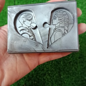 Könnte beinhalten: Eine graue, puzzleförmige Seifenschale mit zwei Herzhälften, jeweils mit dem Profil einer Person. Die Seifenschale wird in einer Hand vor einem grünen Hintergrund gehalten. Das Design hat ein romantisches Thema.