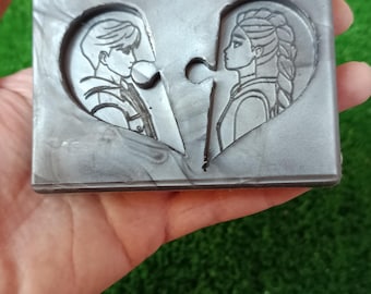 Könnte beinhalten: Eine graue, puzzleförmige Seifenschale mit zwei Herzhälften, jeweils mit dem Profil einer Person. Die Seifenschale wird in einer Hand vor einem grünen Hintergrund gehalten. Das Design hat ein romantisches Thema.