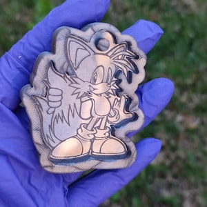 Könnte beinhalten: Ein silberfarbener Schlüsselanhänger mit einem Cartoon-Bild von Tails aus der Videospielserie Sonic the Hedgehog. Der Schlüsselanhänger ist aus einem strukturierten Material gefertigt und hat eine Schlaufe zum Anbringen an einem Schlüsselring.
