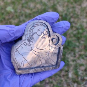 Könnte beinhalten: Ein silberner Schlüsselanhänger mit einem Cartoon-Bild einer Person mit Brille und Schnurrbart. Der Schlüsselanhänger hat die Form eines Personenkopfes.