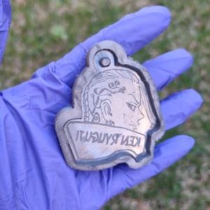 Könnte beinhalten: Ein silberfarbener Schlüsselanhänger aus Metall mit einem detailliert gravierten Bild eines Gesichts und dem Text "KEN RYUGUJI".