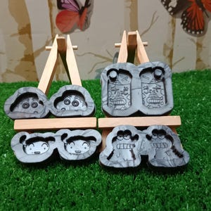 Könnte beinhalten: Vier graue Cartoon-Charakterformen, die auf kleinen Holzstaffeleien ausgestellt sind. Die Formen weisen verschiedene Gesichtsausdrücke auf und sind paarweise angeordnet. Der Hintergrund umfasst eine grüne Oberfläche und eine Kulisse mit Schmetterlingsillustrationen.