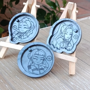 Könnte beinhalten: Drei graue Untersetzer aus Kunstharz mit Cartoon-Illustrationen von Charakteren im Anime-Stil. Jeder Untersetzer hat ein anderes Charakterdesign mit Details wie Frisuren und Kleidung. Die Untersetzer werden auf kleinen Holzstaffeleien ausgestellt.